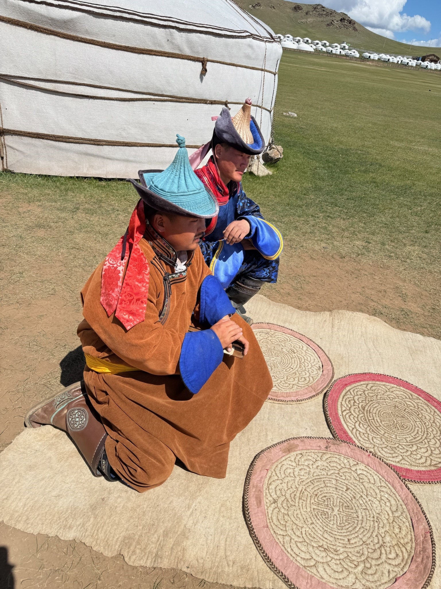 Two men in traditional Mongolian dress seated on felt rugs outside a yurt